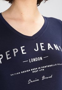 Navy blue T-shirt with a V-neck design, featuring silver printed text: "PEPE JEANS LONDON" and additional brand details. Smooth texture.