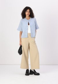 Cardigan bleu clair à manches courtes avec fermeture à boutons, associé à un pantalon large beige et des mocassins noirs, tenant un petit sac noir.