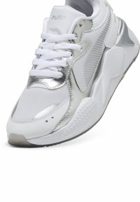 Vit sportskor med silverdetaljer, ovandel i mesh, texturerade sidopaneler och en kraftig sula med ett mönstrat mönster. PUMA-logotypen synlig.