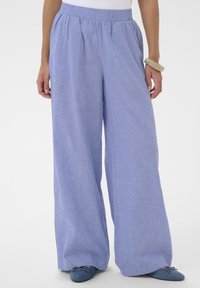 Femme portant un pantalon large violet clair avec une ceinture élastique et des ballerines bleues, debout devant un fond blanc.