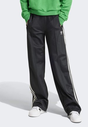 Persoon draagt zwarte Adidas trainingsbroek met witte strepen en witte sneakers, handen in zakken, groene trui zichtbaar vanaf de taille naar beneden.