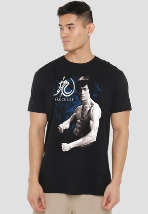 Schwarzes Baumwoll-T-Shirt mit einem grafischen Druck von Bruce Lee in Kampfhaltung, mit blauen Akzenten und stilisiertem Text. Kurze Ärmel.