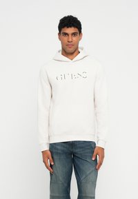 Sudadera con capucha de color beige claro, con un bolsillo frontal y el logo de la marca en relieve. Tejido suave y texturizado con un corte relajado. Combinada con jeans de mezclilla azul.