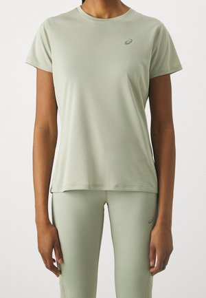 Personne portant un t-shirt de sport vert clair à manches courtes et un legging assorti, debout, bras détendus le long du corps, sur un fond neutre.