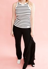 Kvinna i svarta byxor och en vitrandig ärmlös topp står mot en rosa bakgrund, håller en svart jacka i sin högra hand.