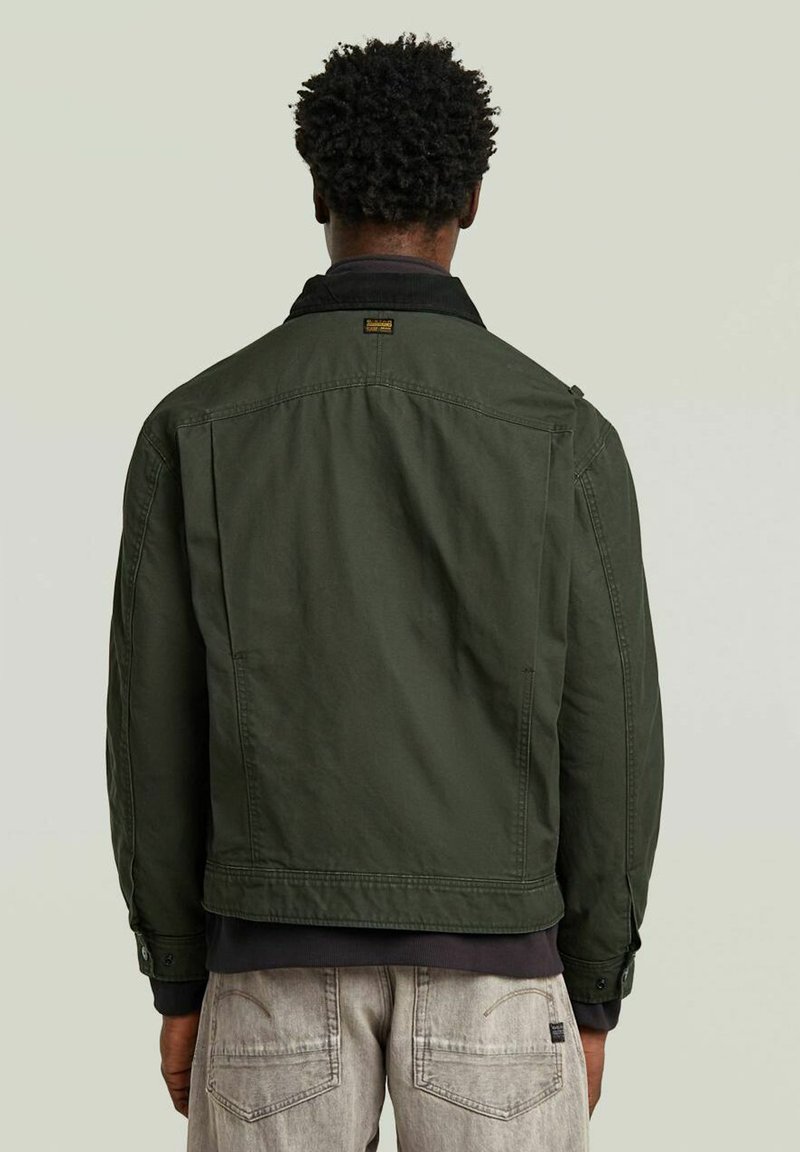 Veste en denim vert olive avec un col noir, un empiècement au dos et deux poches latérales, avec des coutures visibles et une coupe décontractée.