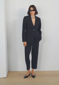 Černý úzký blazer s jedním knoflíkem, přiléhavé kalhoty a černé lodičky na vyšším podpatku. Doplňuje ho metalická řetízková náhrdelník a sluneční brýle.