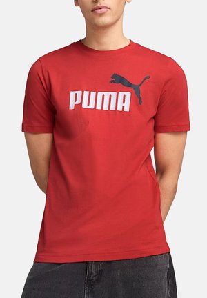 Uomo che indossa una t-shirt rossa Puma con un logo Puma nero e testo Puma bianco, abbinata a pantaloni grigio scuro, in piedi con le mani dietro la schiena.