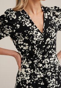Černé zavinovací šaty s květinovým vzorem v bílé barvě. Texturovaná látka s balónovými rukávy a uzlovým detail na pase. Výstřih do V.