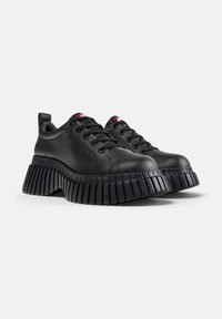 Sneaker in pelle nera con punta arrotondata, suola in gomma spessa e plissettata, e lacci neri. Presenta un cunettino sul tallone e un marchio rosso sulla linguetta.