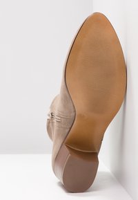 Beżowy zamszowy botek za kostkę z zaokrąglonym noskiem i niskim brązowym obcasem. Podeszwa ma teksturowany grip i widnieje na niej rozmiar 37.