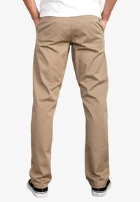 Mand iført beige afslappede bukser og sorte sneakers, stående med hænderne i lommerne, vist fra ryggen mod en hvid baggrund.