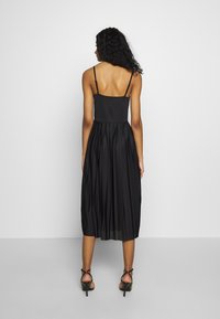 Frau mit langen, lockigen Haaren, die ein schwarzes, plissee Midi-Kleid mit dünnen Trägern und schwarze, geschnürte High Heels trägt, steht vor einer weißen Wand.