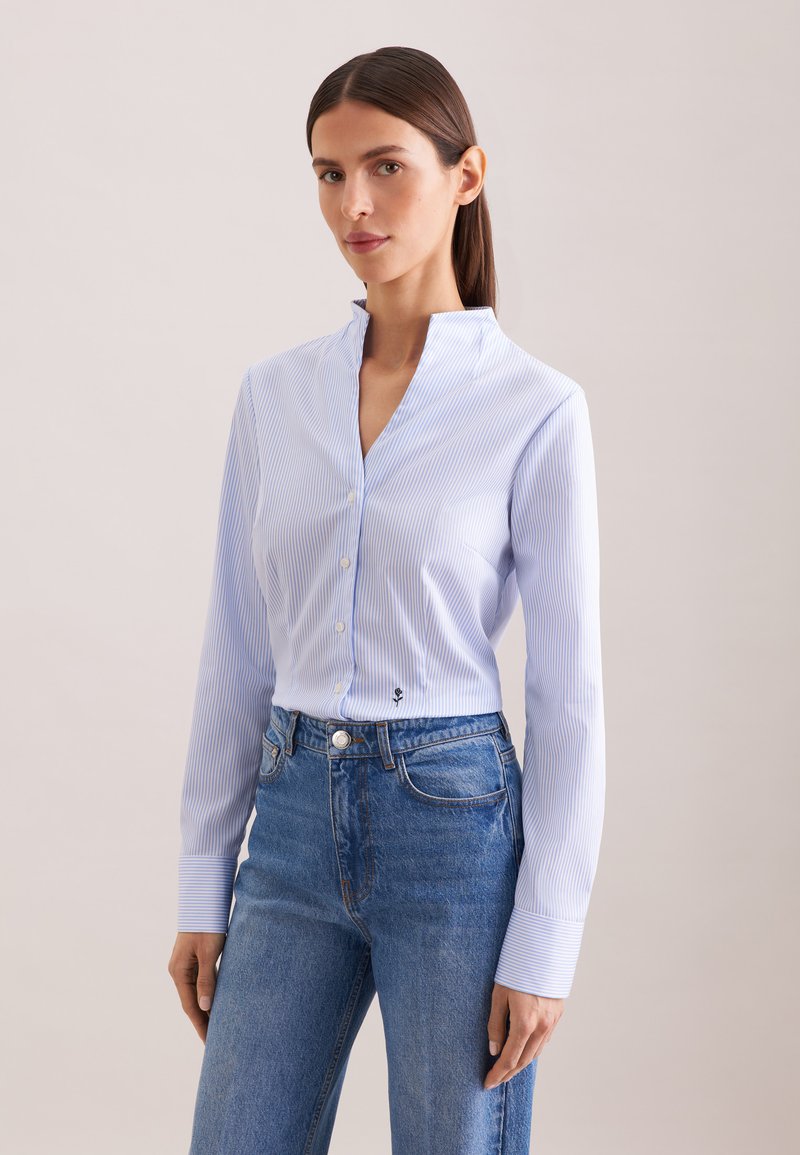 Donna con capelli scuri raccolti in una coda di cavallo, indossa una camicia azzurra a righe sottili con bottoni e jeans blu, su uno sfondo semplice.