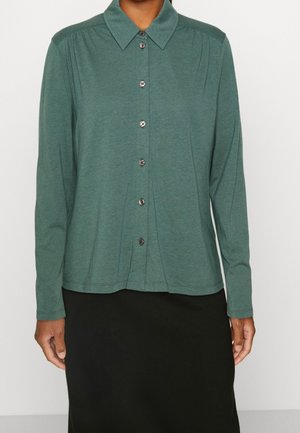 Femme portant une chemise verte à manches longues, boutonnée, avec un col pointu, et une jupe noire.