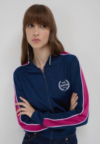 Giacca a zip navy con strisce rosa e bianche sulle maniche, caratterizzata da un logo di Boston e allori bianchi sul petto. Tessuto morbido.