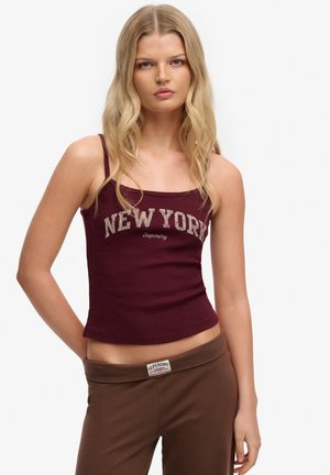 Jeune femme aux longs cheveux blonds portant un débardeur bordeaux "New York" et un pantalon marron avec un logo Superdry visible.