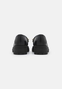 Mocassins en cuir noir avec semelles épaisses à rainures, vus de dos sur fond blanc.