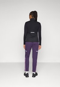 Juodas zip-up ilgomis rankovėmis viršus su tekstūruotu dizainu, derinamas su violetinėmis kelnėmis, turinčiomis baltus akcentus ir šoninius užtrauktukus. Įvaizdį užbaigia juodi batai.