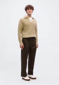 Νεαρός άνδρας με σγουρά μαλλιά, φορώντας ένα μπεζ υφασμάτινο πουλόβερ, σκούρο καφέ παντελόνι, μαύρη ζώνη και λευκά αθλητικά παπούτσια, σε απλό φόντο.