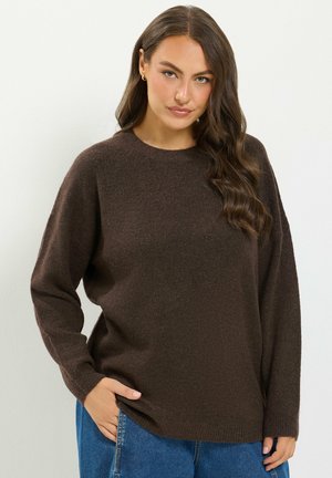 Femme aux longs cheveux bruns ondulés portant un pull marron foncé et un jean bleu, posant avec une main dans la poche devant un fond blanc.