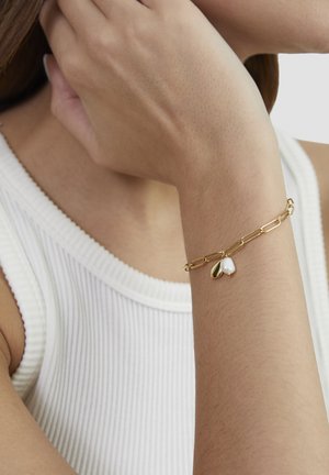 Goldkette-Armband mit zwei Anhängern: ein glatter goldener Tropfen und eine weiße Perle, getragen an einem Handgelenk vor einem strukturierten weißen Oberteil.