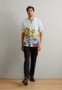 Kortærmet skjorte med knapper i lyseblå med tropisk print, inklusive palmetræer og strandscener. Båret med sorte bukser og sko.