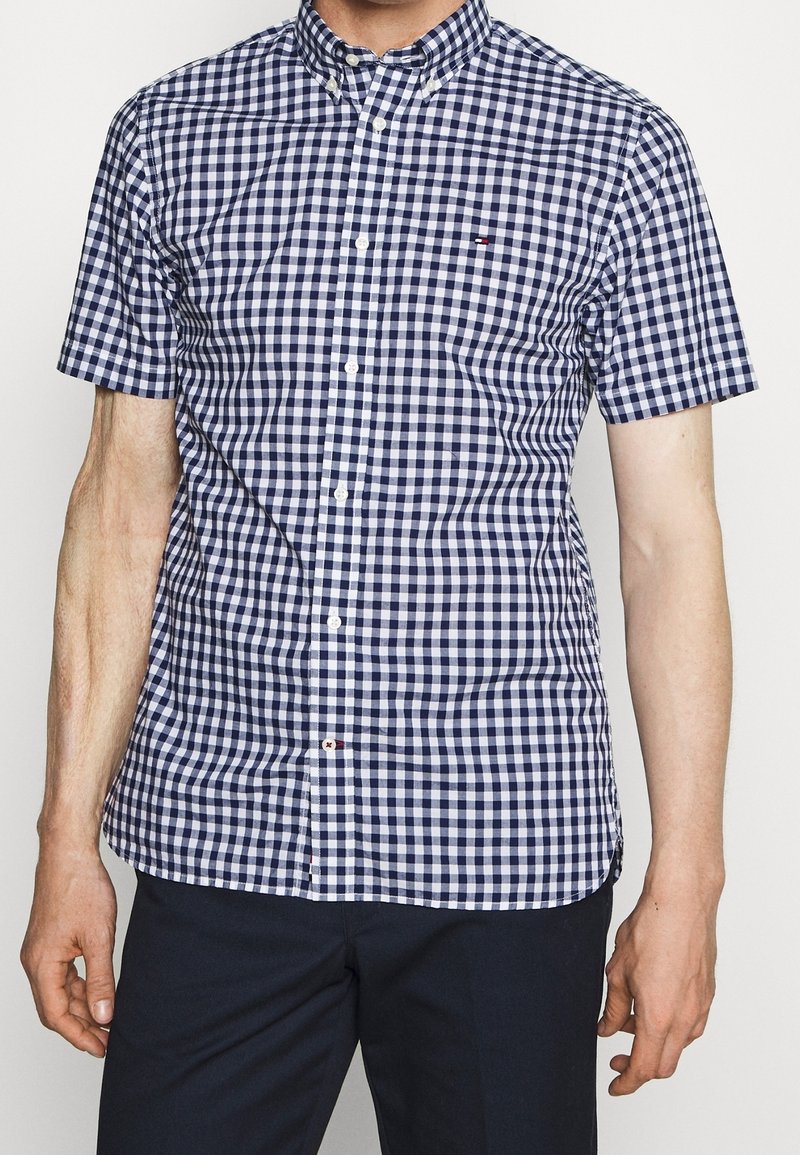 Hombre que lleva una camisa de cuadros en azul marino y blanco de manga corta y pantalones oscuros, de pie contra un fondo liso.