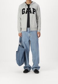 Grauer Kapuzenpullover mit Reißverschluss und schwarzer "G.A.P."-Aufschrift über einem schwarzen T-Shirt. Locker sitzende blaue Jeans und eine blaue Canvas-Tragetasche. Schwarze Schuhe.