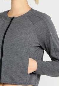 Grå, kortärmad träningsjacka med en texturerad yta, dragkedjestängning och sidofickor. Har raglanärmar och rund halsringning.