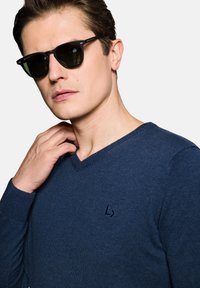 Granatowy sweter V-neck z dzianiny, z małym logo marki na piersi i ściągaczami przy rękawach. Czarne okulary przeciwsłoneczne z przyciemnionymi soczewkami.