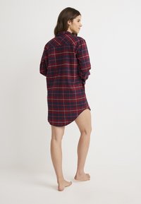 Camisa de franela a cuadros rojos y marinos con mangas largas, cuello y un dobladillo curvado. La parte trasera cuenta con un yugo y aberturas laterales para un ajuste relajado.