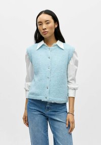 Lyseblå strikket vest med v-hals, ermeløs design og sølvfargede knapper, lagdelt over en hvit skjorte med krage.