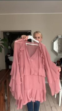 Vestido rosa de capas con mangas y dobladillo con volantes, confeccionado en tela ligera. Diseño de escote en V y silueta fluida, colgado en una percha.