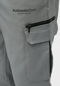 Szürke nadrág sima textúrával, cipzáras zsebbel, varrott díszítésekkel és "AuthenticDept." márkajelzéssel.