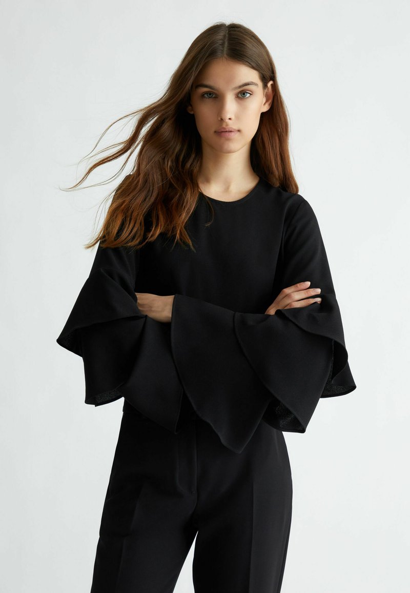 Top nero con ampie maniche a campana e scollatura rotonda, realizzato in tessuto morbido. La modella posa con le braccia incrociate su uno sfondo semplice.