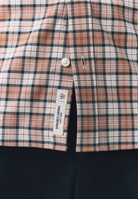 Chemise en coton à carreaux avec un mélange de carrés pêche, marine et blanc, col arrondi, fermeture par boutons, et une étiquette avec le texte "DURABLE GOODS".