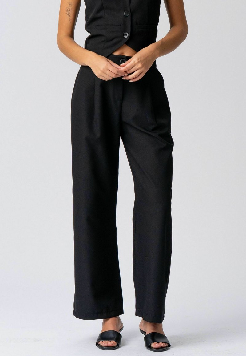 Femme portant un pantalon noir ample taille haute, un gilet noir sans manches à boutons et des sandales plates noires à bouts ouverts, debout devant un fond uni.