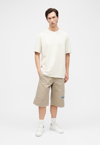 Jeune homme debout portant un t-shirt surdimensionné blanc cassé, un short beige mi-genou avec une poche à rabat, des chaussettes blanches et des baskets blanches.