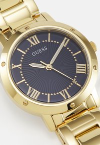 Montre en acier inoxydable doré avec un cadran bleu marine, des index en chiffres romains et un motif texturé. Elle présente des aiguilles pour les heures, les minutes et les secondes.