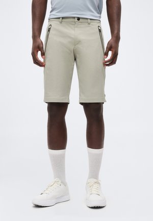 Man wearing light beige knee-length shorts with side zipper pockets, white crew socks, and white sneakers standing against a plain background.