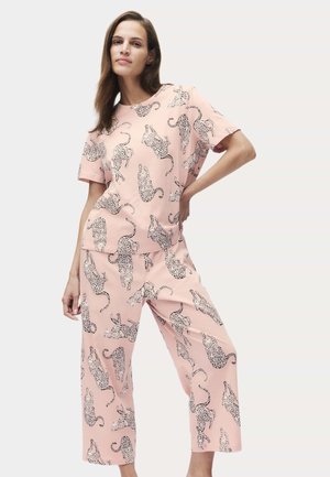 Kvinde iført pink top med korte ærmer og vidde bukser med sort-hvidt leopardprint, poserer med hånden på hoften mod en lys baggrund.