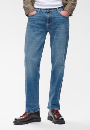 Person trägt blaue Jeans, braune Lederstiefel mit roten Schnürsenkeln, ein weißes Hemd und eine braun karierte Jacke mit Händen in den Taschen.
