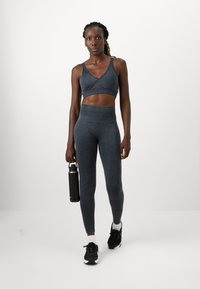 Atletska žena u tamnosivom sportskom kompletu i crnim tenisicama drži crnu plastičnu bocu za vodu, krećući se prema naprijed na jednostavnoj bijeloj pozadini.