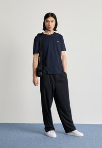 Jūrininkų crew-neck marškinėliai su mažomis baltomis logotipu, derinami su juodais smulkiais dryžiais kelnais ir baltais sportbačiais. Plonas dizainas, kasdieniška medžiaga, laisvas fitas.