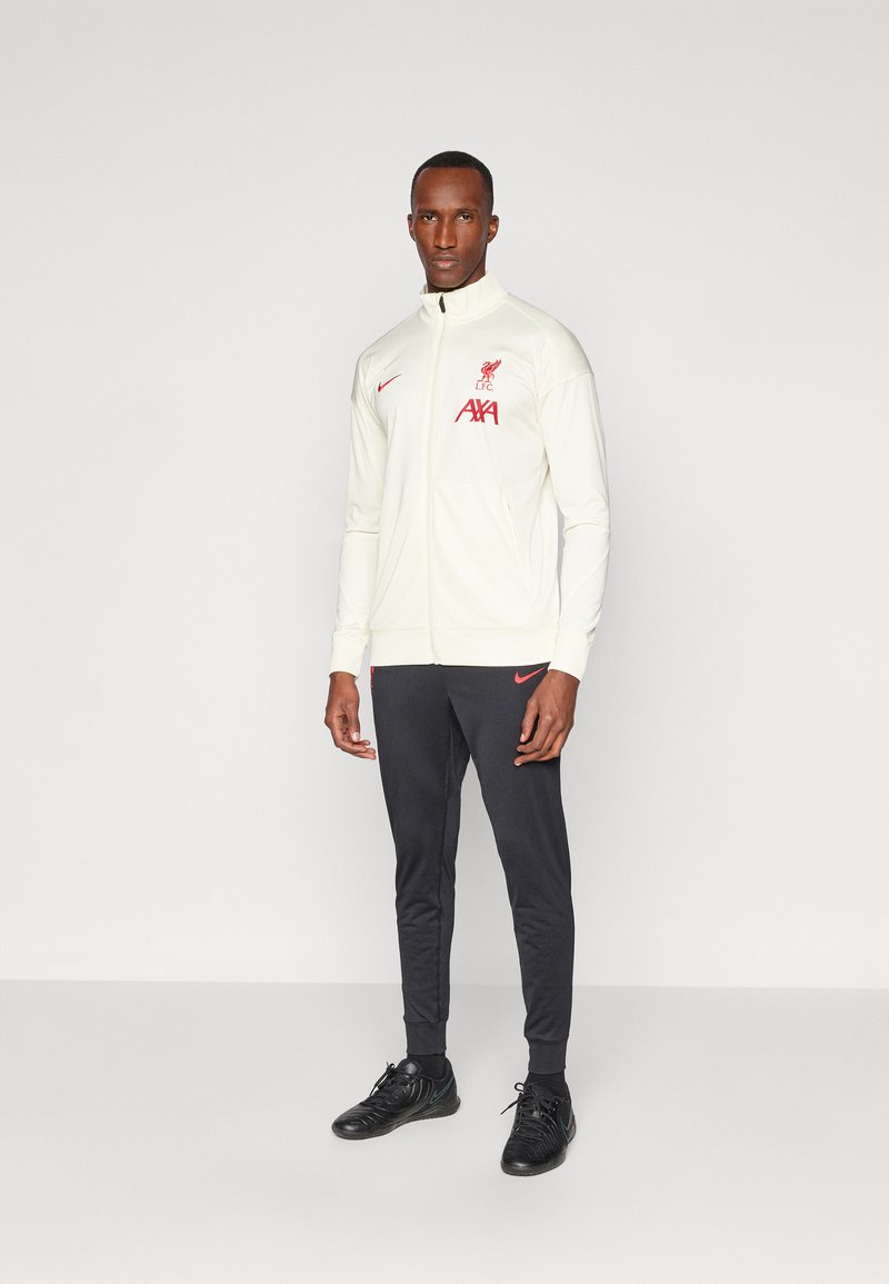 Krem jakna na zip s crvenim logom Liverpool FC, crne hlače i crne sportske cipele. Glatka tkanina, prilagođeni dizajn i džepovi na zatvarač.
