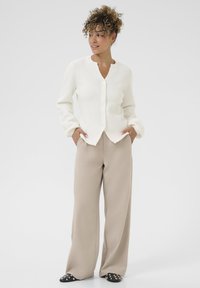 Femeie cu păr creț poartă un cardigan alb cu nasturi, pantaloni bej cu croială largă și pantofi negri cu tinte, stând cu mâinile în buzunare.