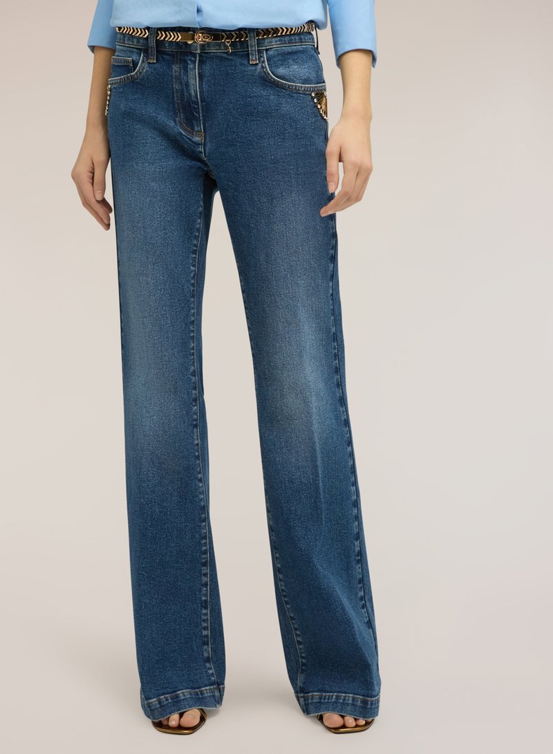 Frau trägt weit geschnittene blaue Jeans mit verzierten vorderen Taschen, einen gemusterten Gürtel, ein blaues Langarmshirt und offene Sandalen.