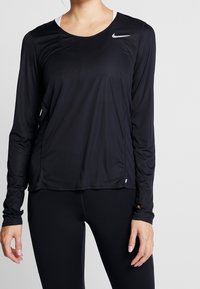 Mustapaita, jossa on pitkät hihat, valmistettu kevyestä kankaasta, pyöreä kaula-aukko ja pieni valkoinen logo vasemmassa olkapäässä.