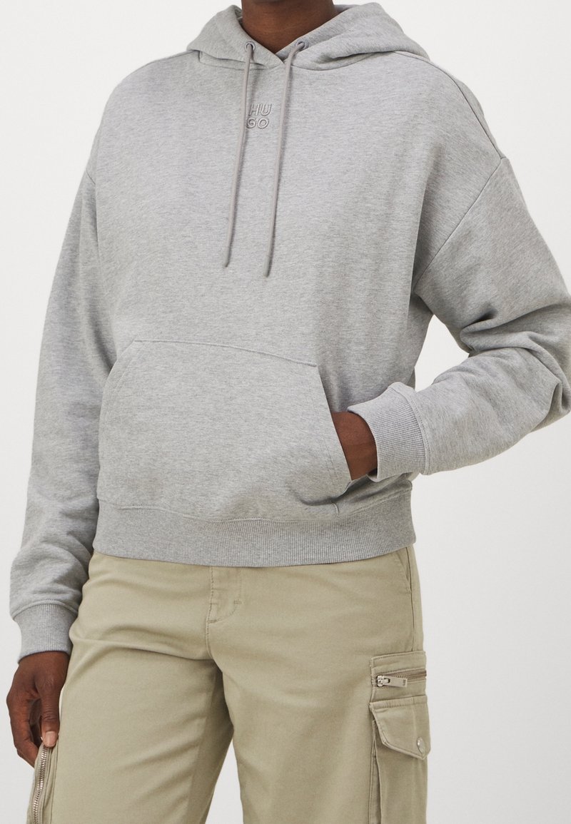 Personne portant un sweat à capuche gris clair avec poche avant et un pantalon cargo beige avec poche zippée, sur un fond blanc uni.